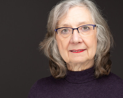 Business headshot of a female with grey hair and glasses, wearing a high necked purple top, shot against a dark grey background